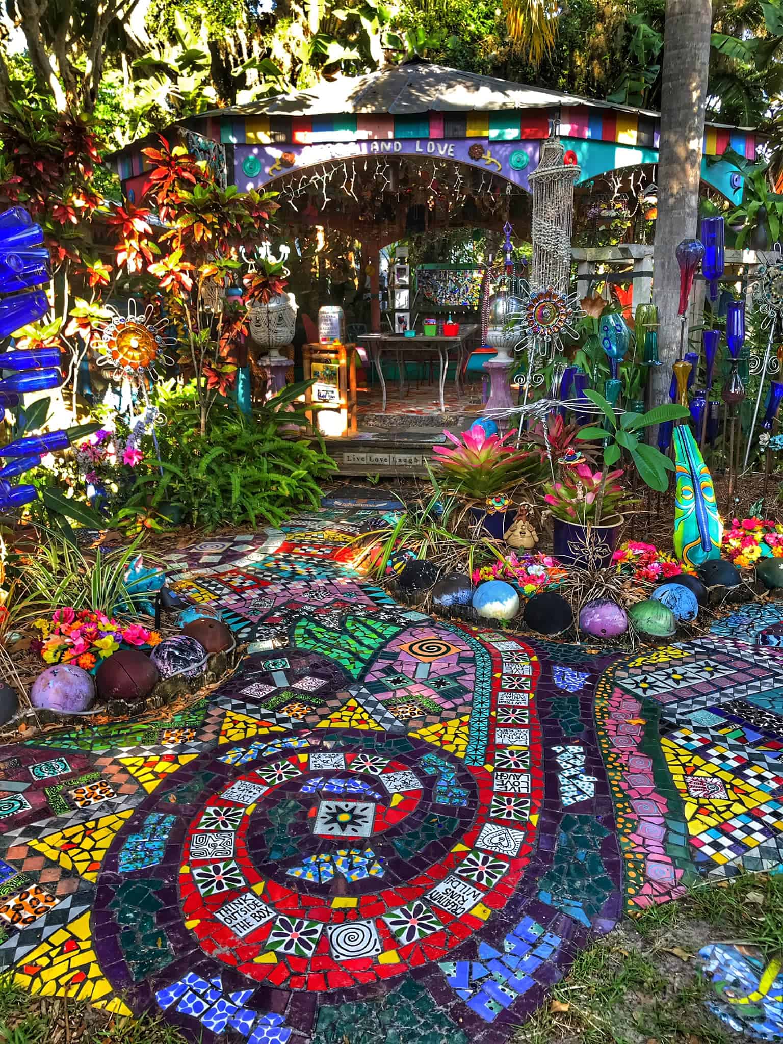Outdoor portion of Whimzeyland. Brightly colored tiles make a swirl path, lots of ornate sculptures, bowling balls, and other piece hang from trees and are covering the ground