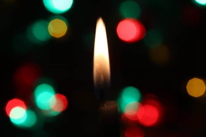 Single candle flame in foreground, with bright, colorful lights blurred behind it