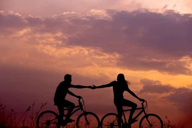 Couple riding bikes at sunset, reaching arms towards each other