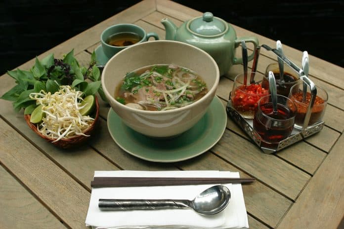 Bowl of Pho on table, with bean sprouts, basil, and sauces surrounding the bowl