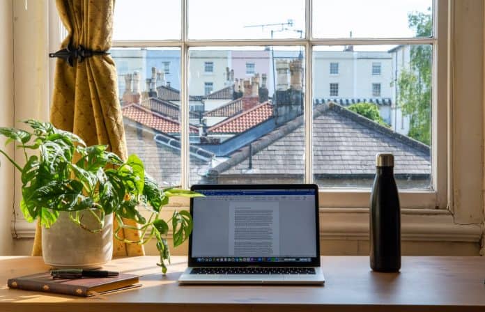 Desk in front of sunny window, overlooking other homes, with laptop, plant, and waterbottle on desk