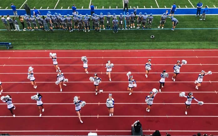 Group of cheerleaders on track, doing a routine, while football players stand on the sideline