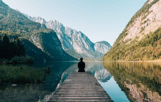 Man sitting at the end of a dock on a lake, between mountains